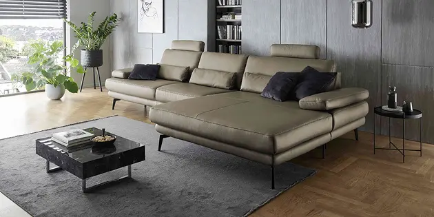 Stilvolles Wohnzimmer mit großem olivgrünem Ledersofa und breiter Recamiere auf schwarzen Metallfüßen. Dunkle Akzentwand mit Bücherregal, moderner Couchtisch auf Teppich, Pflanzen am Fenster und warmes Parkett sorgen für elegante Atmosphäre.